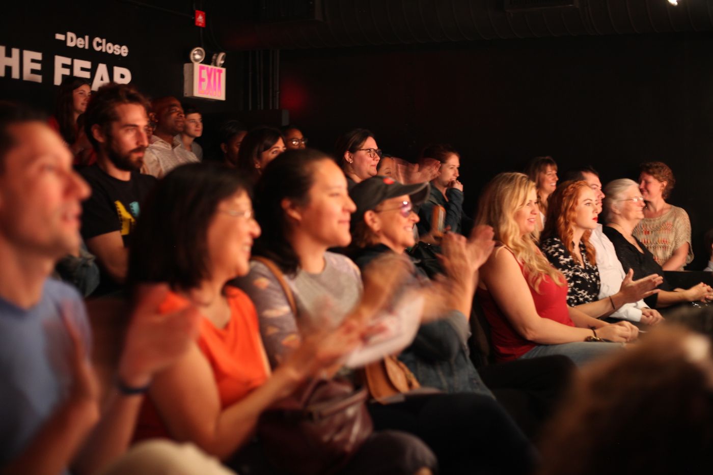 A packed audience in a small theater laughs and applauds.