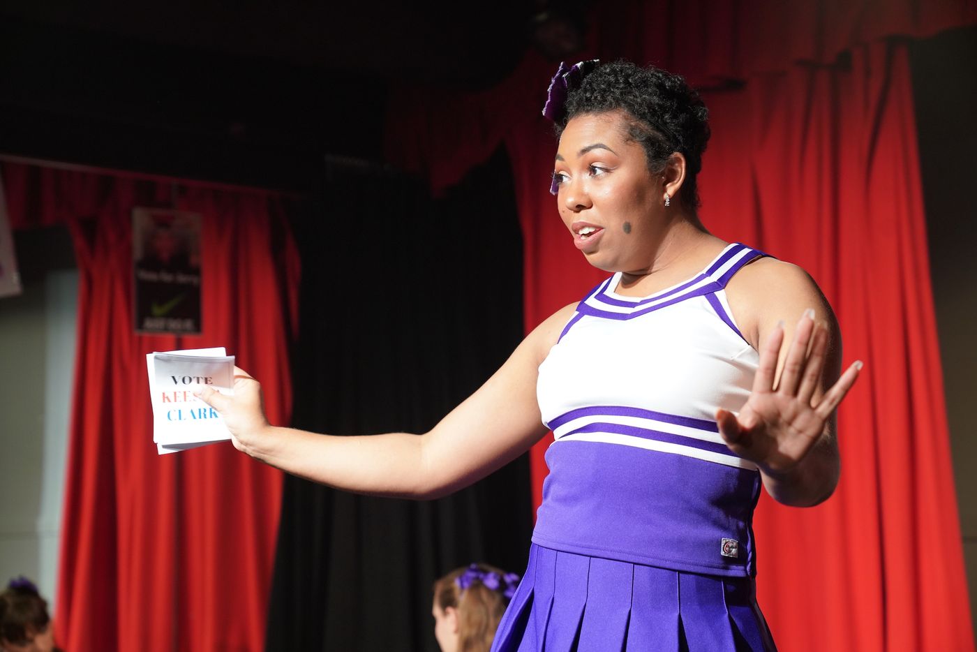 Keesha, in a cheer uniform, holds out a campaign flyer and raises her other hand on stage.