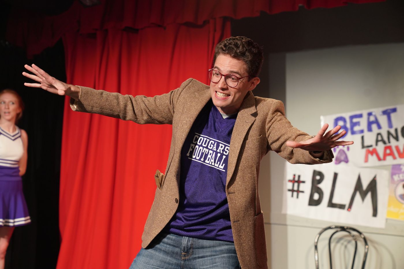 An actor in a brown blazer and a Douglas Football shirt throws his arms wide in front of a red curtain and a #BLM sign.