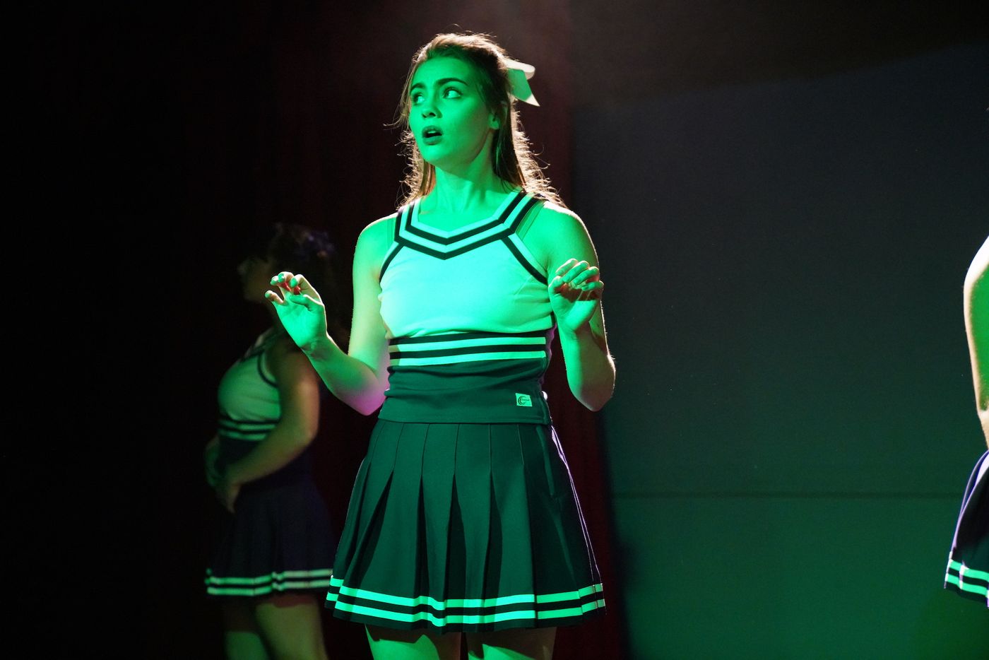 A cheerleader in a purple and white uniform, lit green on a dark stage, looks out in alarm.