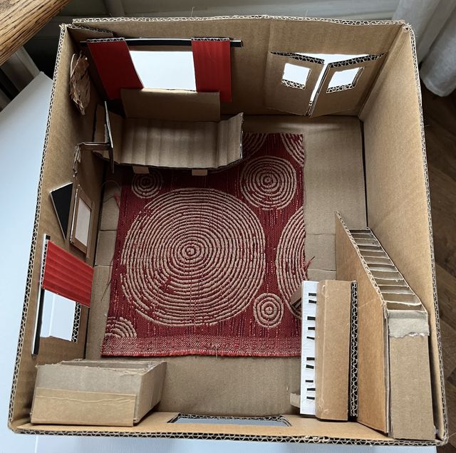 Cardboard scale model of a living room seen from above, with a red patterned rug, windows with red curtains and small cardboard furniture.