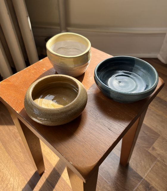 Three small wheel-thrown bowls on a wooden stool in sunlight: one cream, one speckled olive, one glossy dark blue.